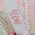 A hand holds two decorative bookmarks in front of an open book. One bookmark is light pink with pink script text reading 'and if the music is good you DANCE', and the other has a repeating pink ribbon and bow pattern on a white background.
