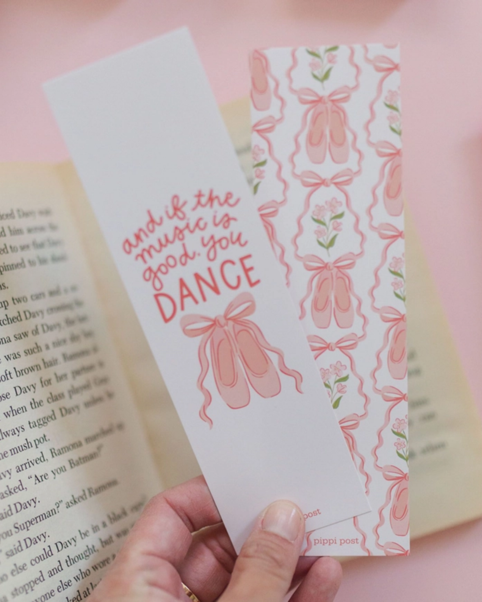 A hand holds two decorative bookmarks in front of an open book. One bookmark is light pink with pink script text reading 'and if the music is good you DANCE', and the other has a repeating pink ribbon and bow pattern on a white background.
