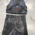A two-piece activewear set consisting of a sports bra and matching shorts, both featuring a dark gray and black crocodile-embossed pattern with a small pink logo on the front. The set is laid flat on a white marble background.