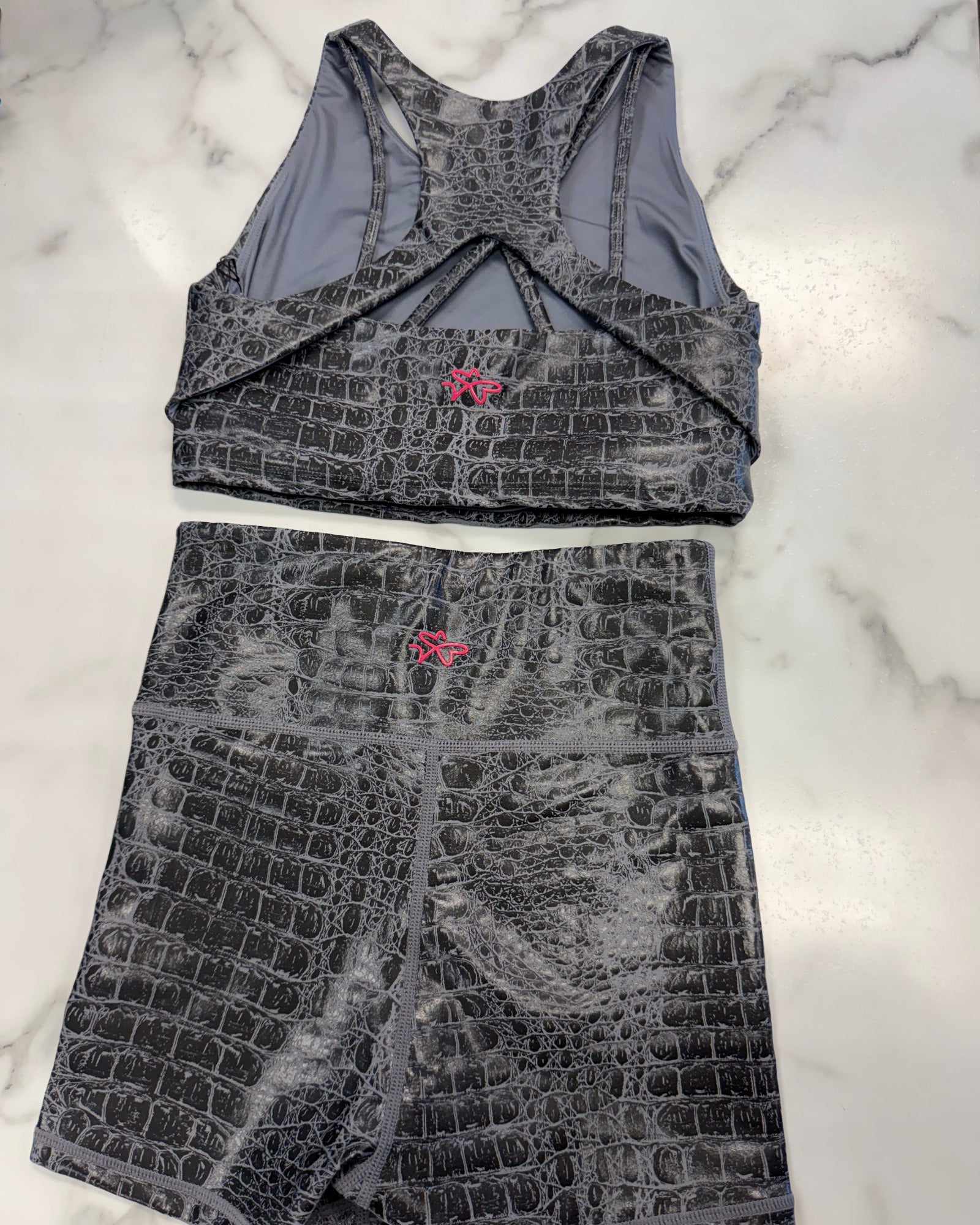 A two-piece activewear set consisting of a sports bra and matching shorts, both featuring a dark gray and black crocodile-embossed pattern with a small pink logo on the front. The set is laid flat on a white marble background.