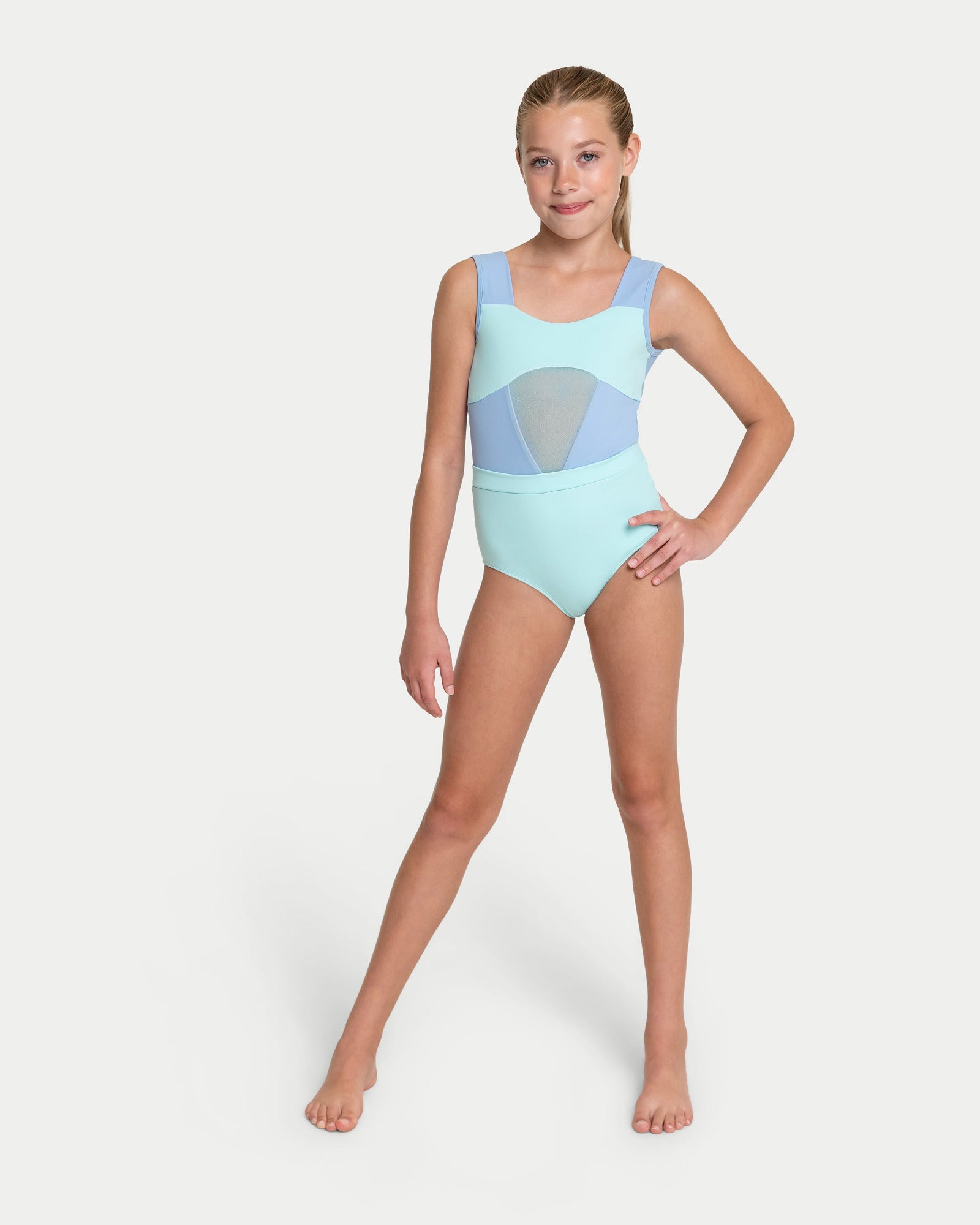 A young girl modeling a light blue leotard with a light mint green and light blue geometric pattern on the chest, standing against a solid light gray background. 