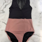 A color-blocked leotard laid flat on a white marble background. The top half is solid black with a sheer mesh V-neck detail, and the bottom half is a dusty rose pink.