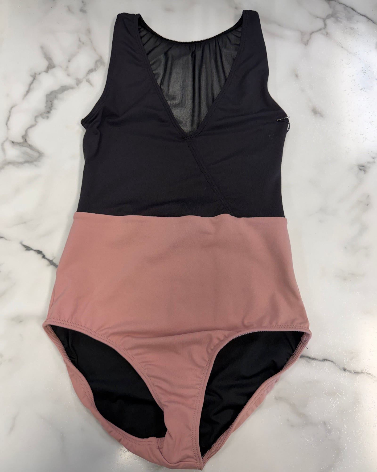 A color-blocked leotard laid flat on a white marble background. The top half is solid black with a sheer mesh V-neck detail, and the bottom half is a dusty rose pink.