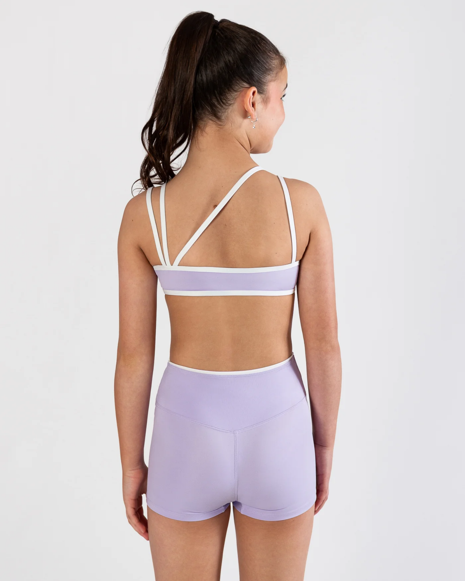 A woman shown from the back wearing a lavender and white two-piece dance set, consisting of a sports bra with crisscross straps and matching high-waisted shorts, against a plain white studio background.