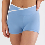 A close-up of a model wearing light blue dance shorts with a white crossover waistband detail, shown against a plain light gray studio background.