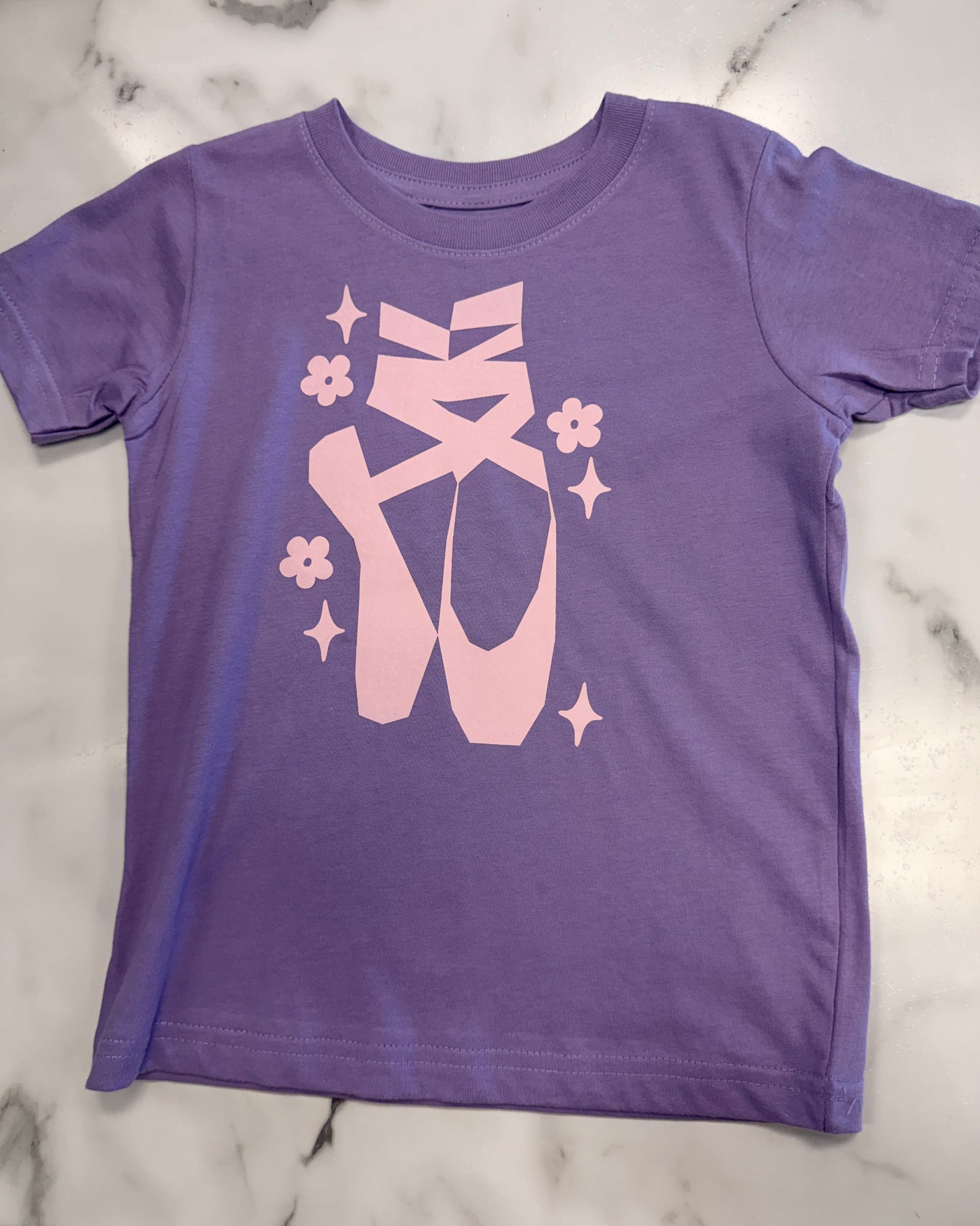 A purple short-sleeve t-shirt with a pink graphic of ballet pointe shoes, ribbon bows, and small flowers, laid flat on a white marble surface.