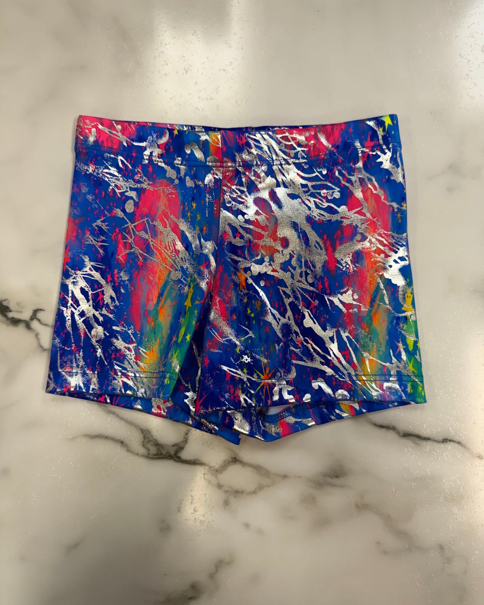 A pair of high-waisted dance shorts featuring a vibrant, multicolor paint splatter pattern on a shiny blue base, laid flat on a white marble surface.