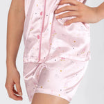 A close-up of a person wearing a light pink satin pajama set with a whimsical ballet shoe and star print, featuring a zippered top and matching shorts.