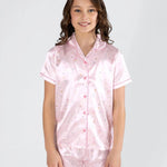 A young girl with wavy brown hair stands against a light gray studio background wearing a light pink satin pajama set with a small multicolor pattern. The set includes a short-sleeve button-front pajama top and matching shorts.