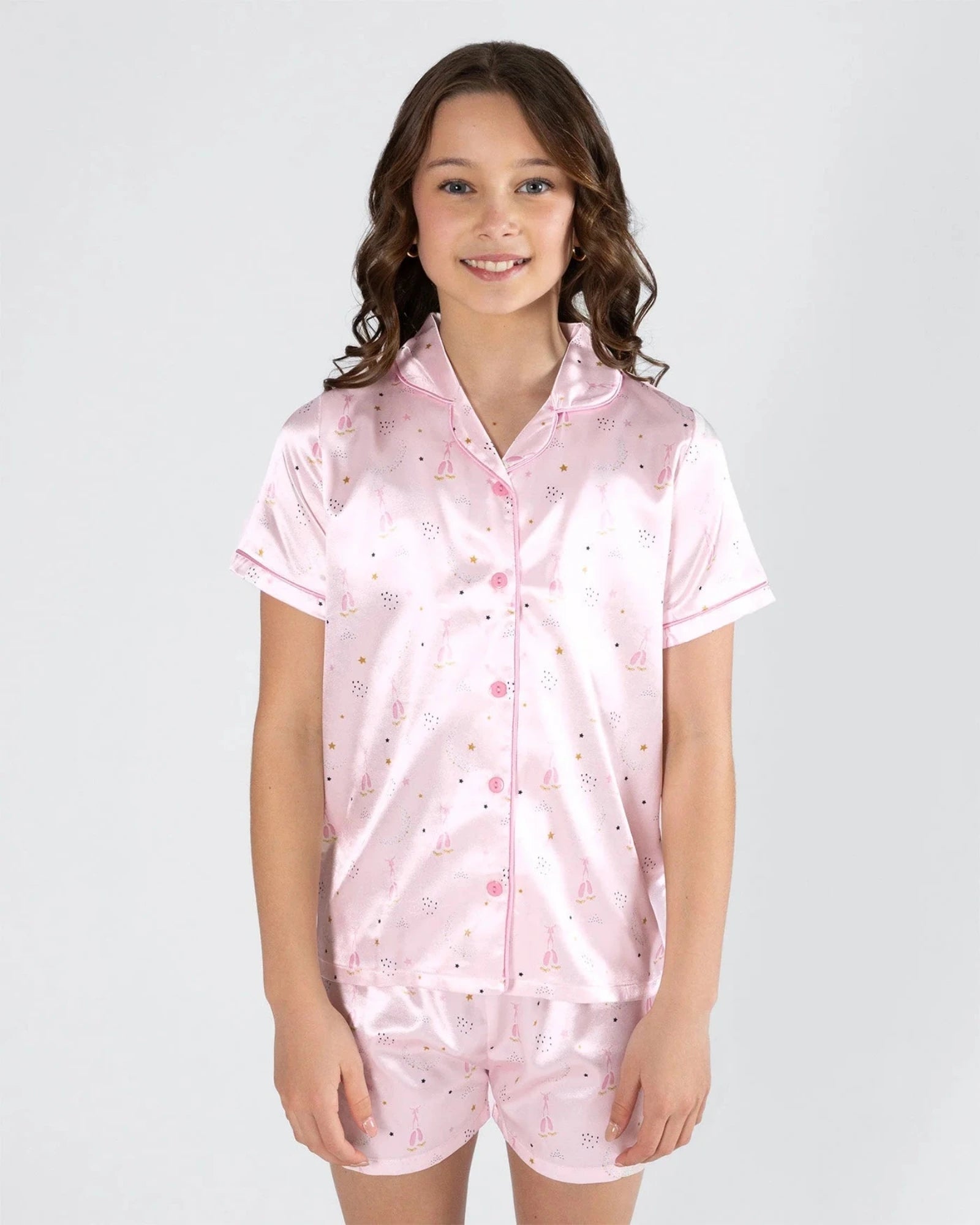 A young girl with wavy brown hair stands against a light gray studio background wearing a light pink satin pajama set with a small multicolor pattern. The set includes a short-sleeve button-front pajama top and matching shorts.