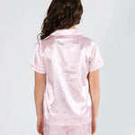 A woman viewed from the back wearing a light pink satin pajama set with a small multicolor ballet and star print, consisting of a short-sleeve collared top and matching shorts.