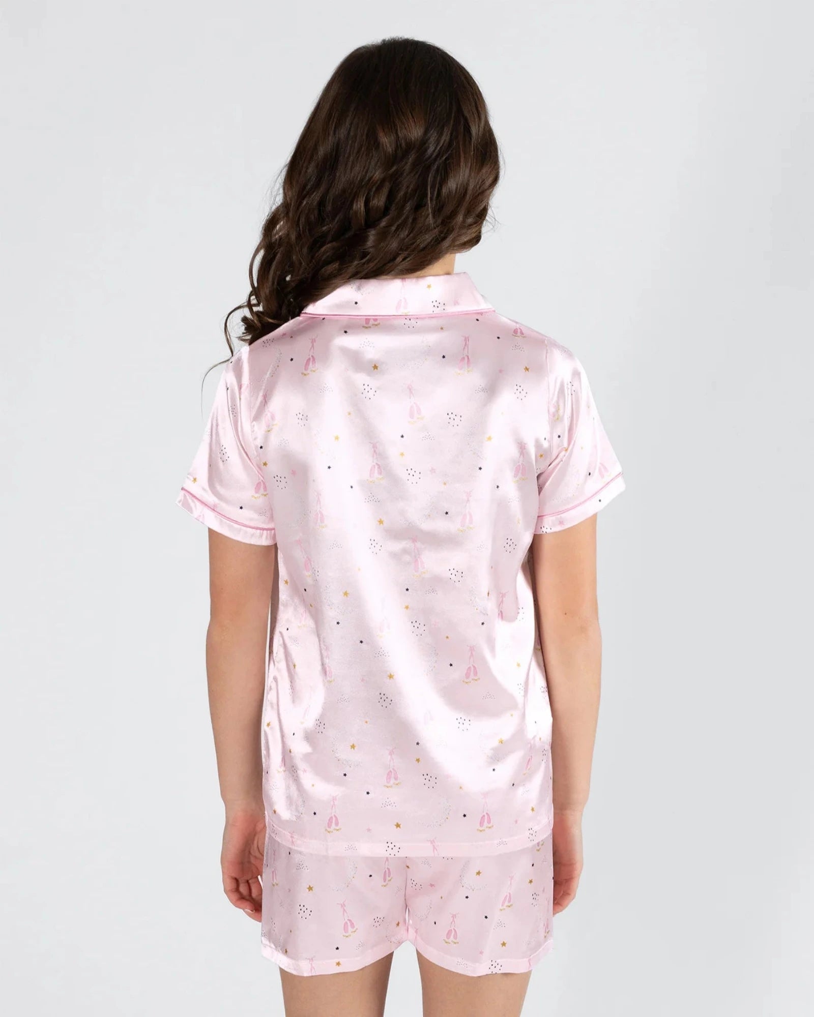 A woman viewed from the back wearing a light pink satin pajama set with a small multicolor ballet and star print, consisting of a short-sleeve collared top and matching shorts.