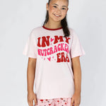 A young girl smiling while wearing a light pink graphic pajama set with red lettering that reads 'IN MY NUTCRACKER ERA' and matching pink patterned shorts, set against a plain light gray background.