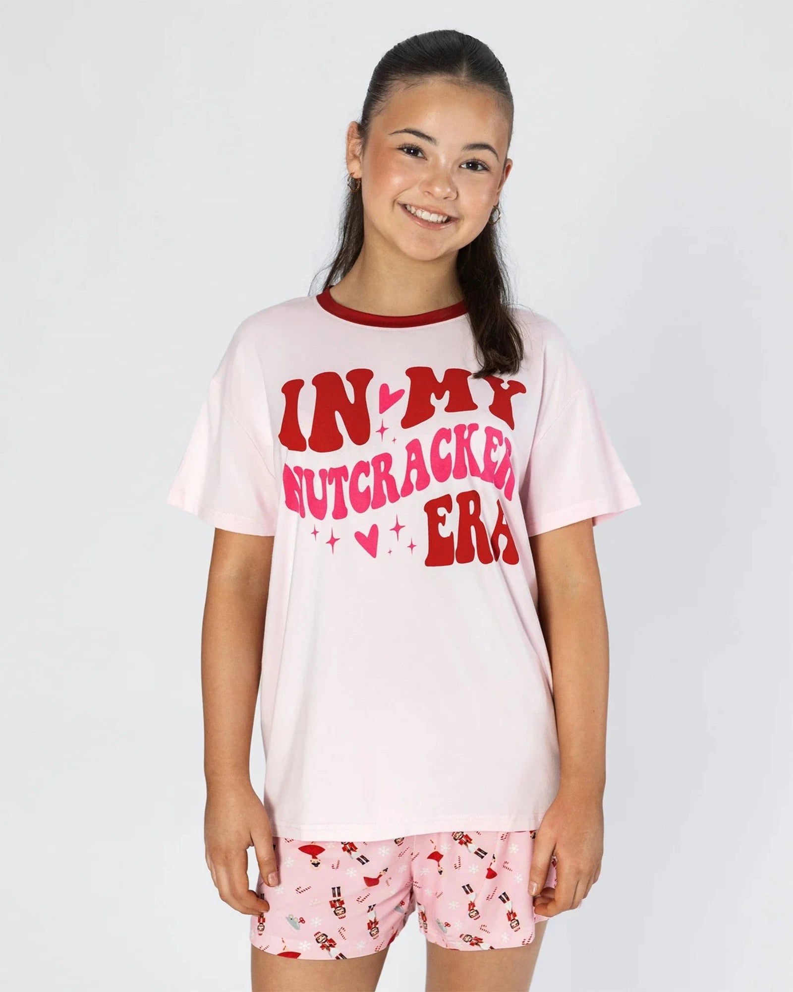 A young girl smiling while wearing a light pink graphic pajama set with red lettering that reads 'IN MY NUTCRACKER ERA' and matching pink patterned shorts, set against a plain light gray background.