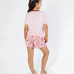 A young girl shown from the back wearing a light pink short-sleeve pajama top with a red collar and matching pink pajama shorts covered in a small red heart pattern. She is standing against a clean white studio background.