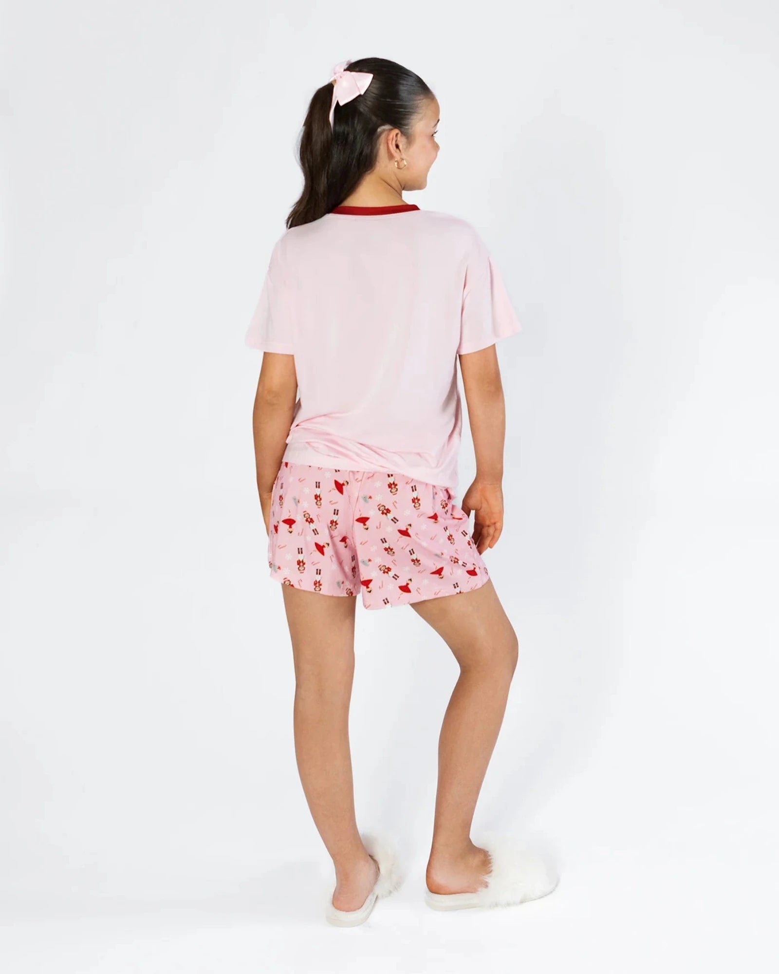 A young girl shown from the back wearing a light pink short-sleeve pajama top with a red collar and matching pink pajama shorts covered in a small red heart pattern. She is standing against a clean white studio background.
