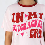 A close-up of a person wearing a light pink ringer t-shirt with a red crew neck collar and a bold graphic print that reads 'IN MY NUTCRACKED ERA' in red and hot pink lettering with small heart and star accents.