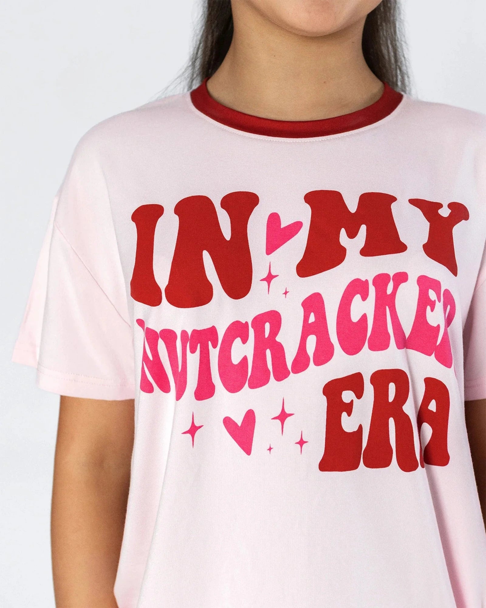 A close-up of a person wearing a light pink ringer t-shirt with a red crew neck collar and a bold graphic print that reads 'IN MY NUTCRACKED ERA' in red and hot pink lettering with small heart and star accents.