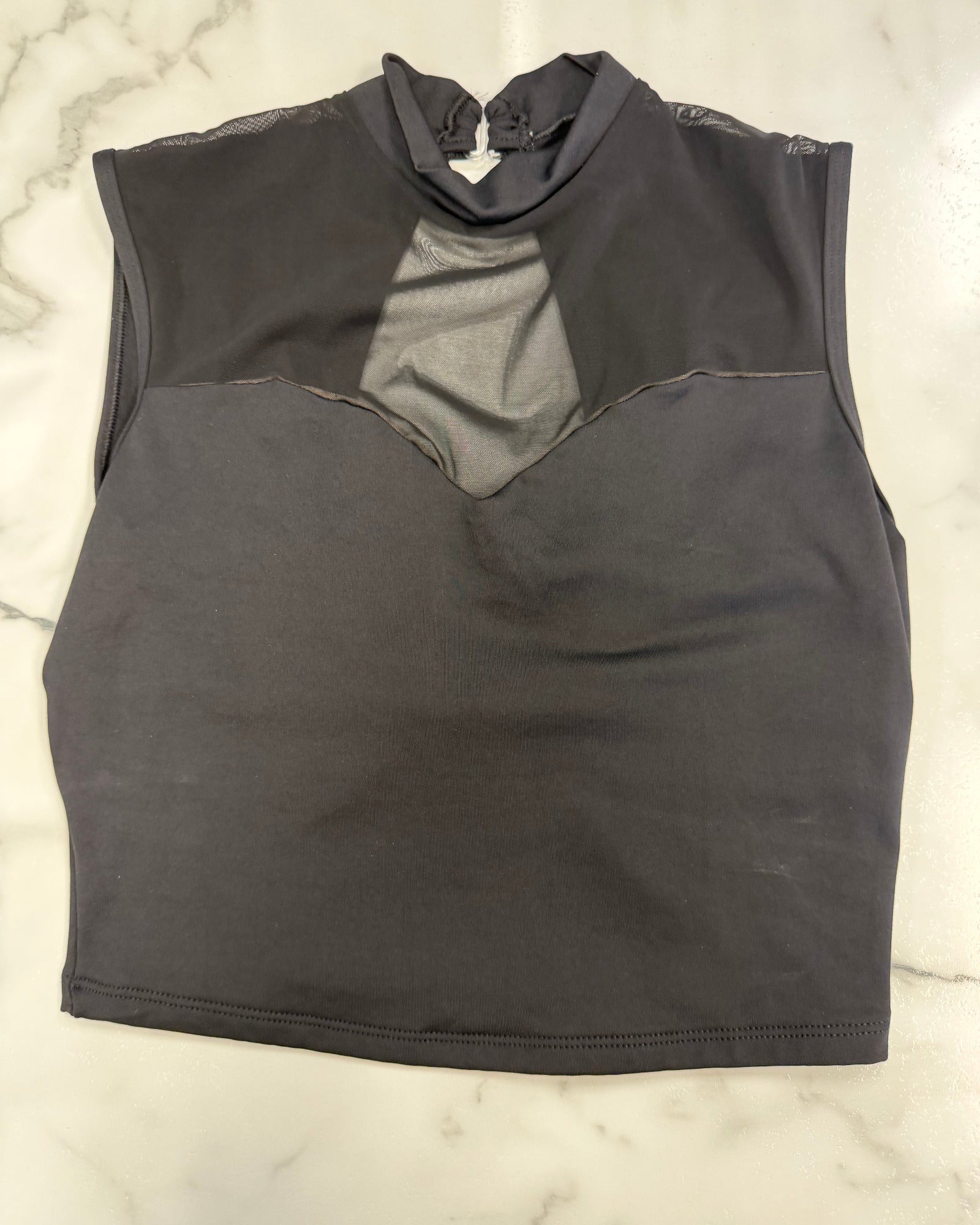 A dark black sleeveless mock neck top with a sheer mesh yoke, laid flat on a white marble surface.