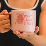 A person in a black tank top holds a light pink ceramic mug with a pink ballet shoe graphic and the text 'POINT YOUR TOES' on the front.