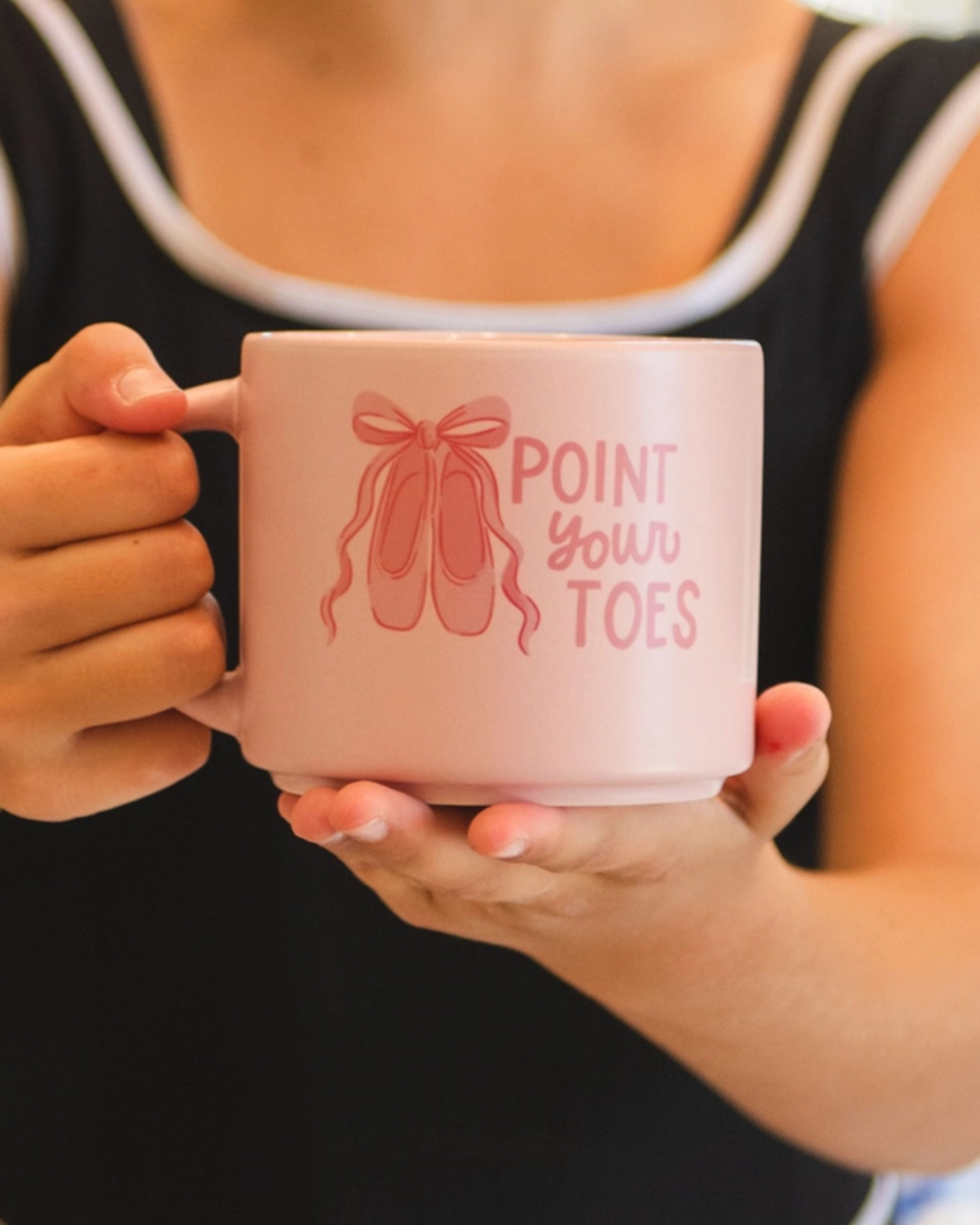 A person in a black tank top holds a light pink ceramic mug with a pink ballet shoe graphic and the text 'POINT YOUR TOES' on the front.