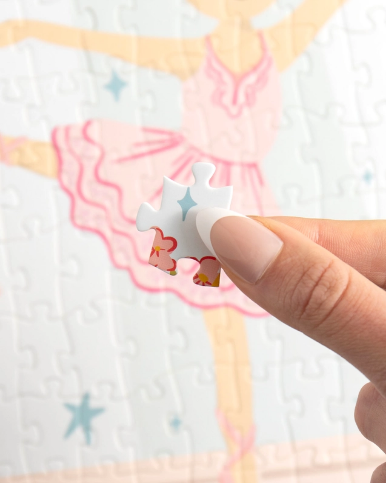 A close-up of a hand holding a white jigsaw puzzle piece with a blue star and small orange flower, with a partially completed pastel ballerina puzzle in the blurred background.
