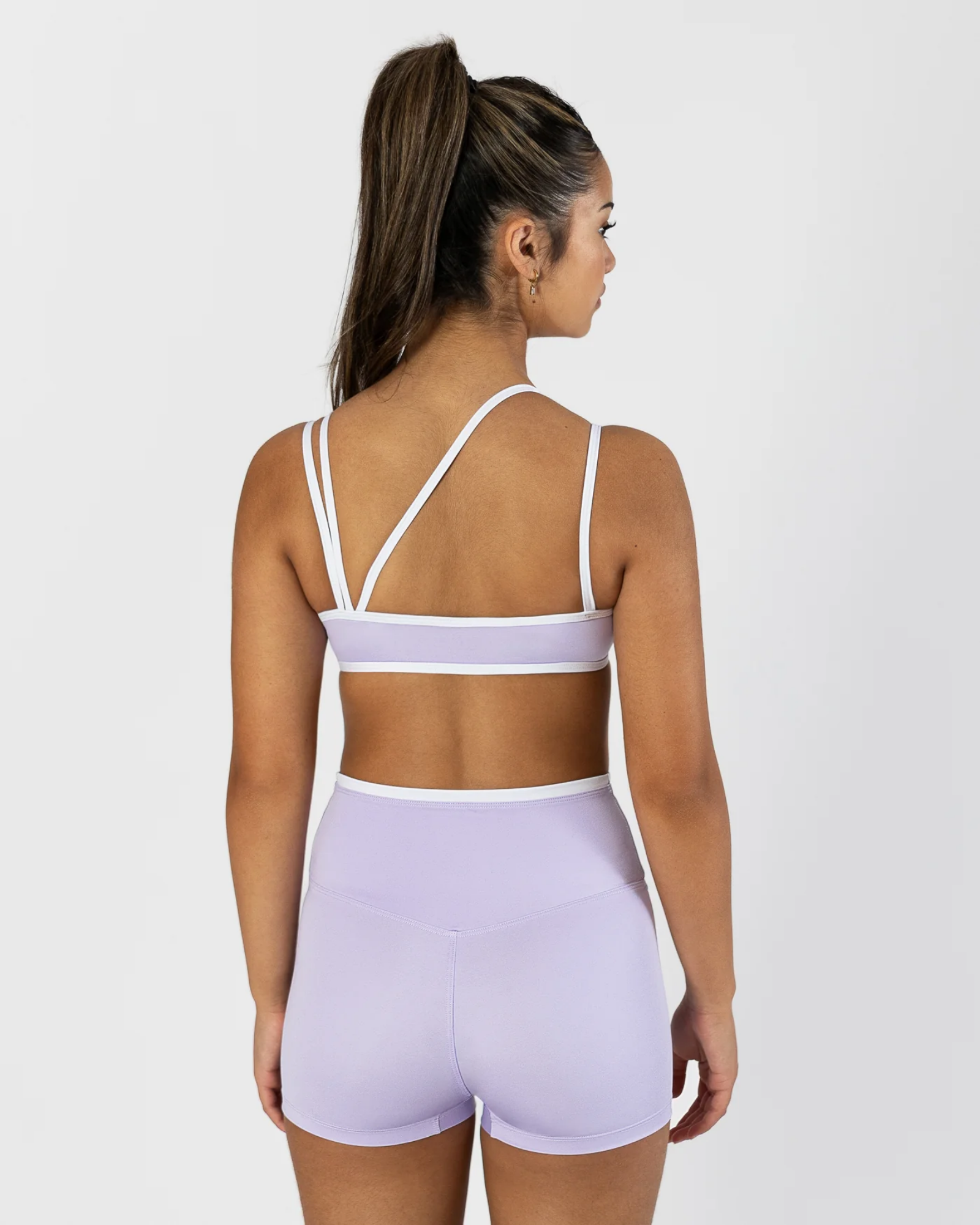 A woman shown from the back wearing a light lavender two-piece dance set with white trim, consisting of a sports bra and high-waisted shorts, against a plain light gray background.