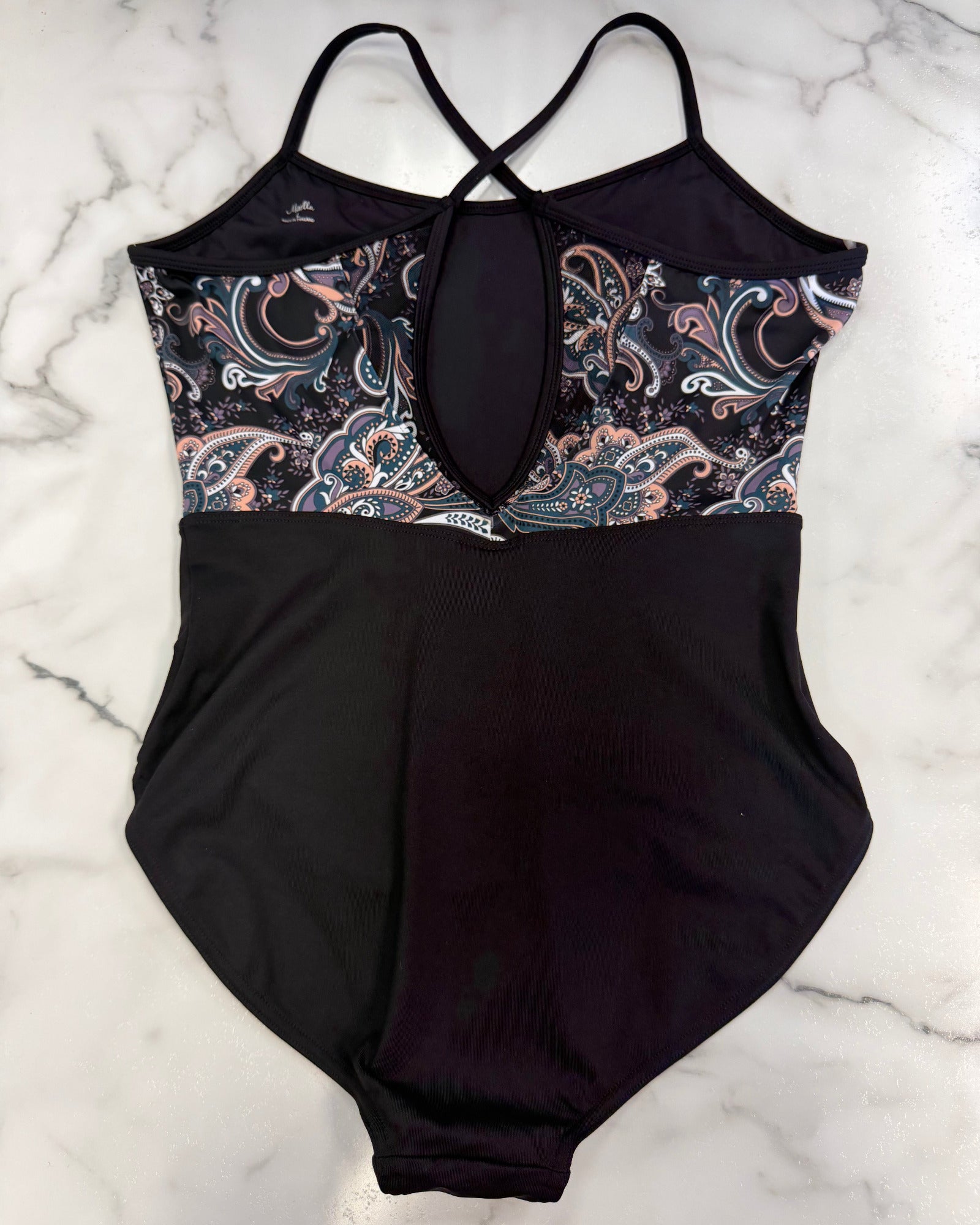 A leotard laid flat on a white marble surface. The bottom half is solid black, while the upper bust and underwire area feature a paisley pattern in black, dusty pink, and white.