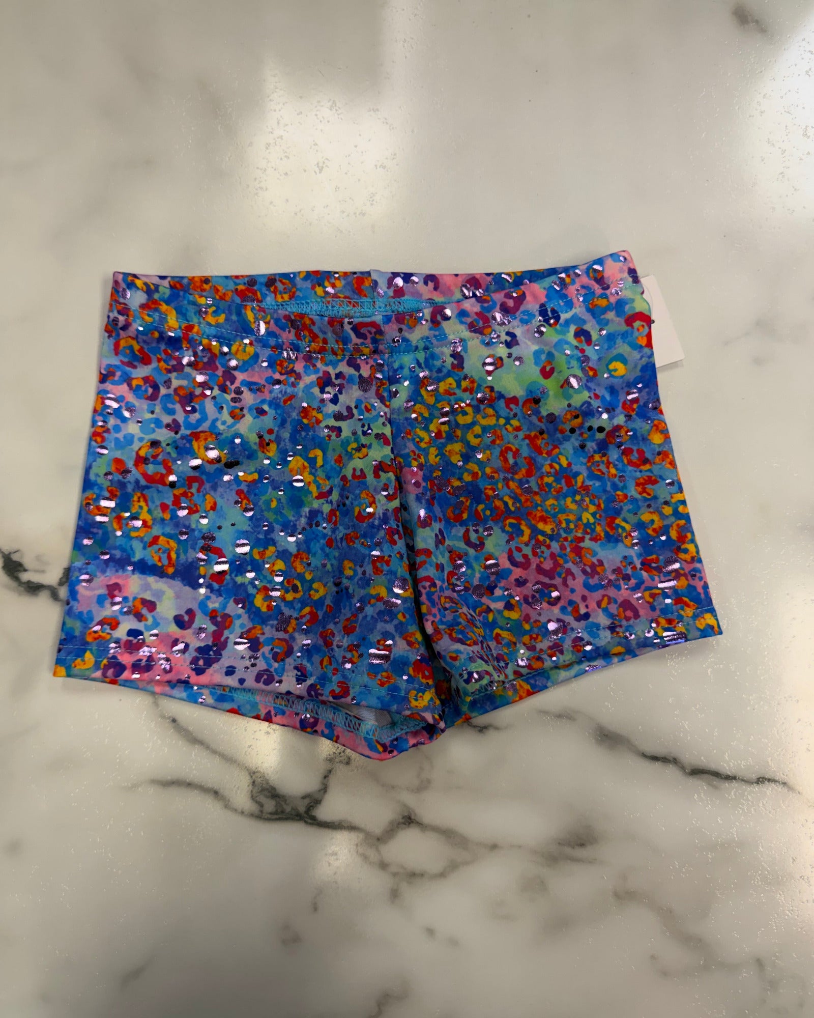 A pair of colorful leopard-print swim trunks with sequins, laid flat on a white marble surface. The pattern features a mix of blue, pink, orange, yellow, and red.