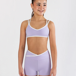 A young girl modeling a lavender athletic two-piece set consisting of a dance bra and matching high-waisted shorts with white trim, set against a plain light gray background.