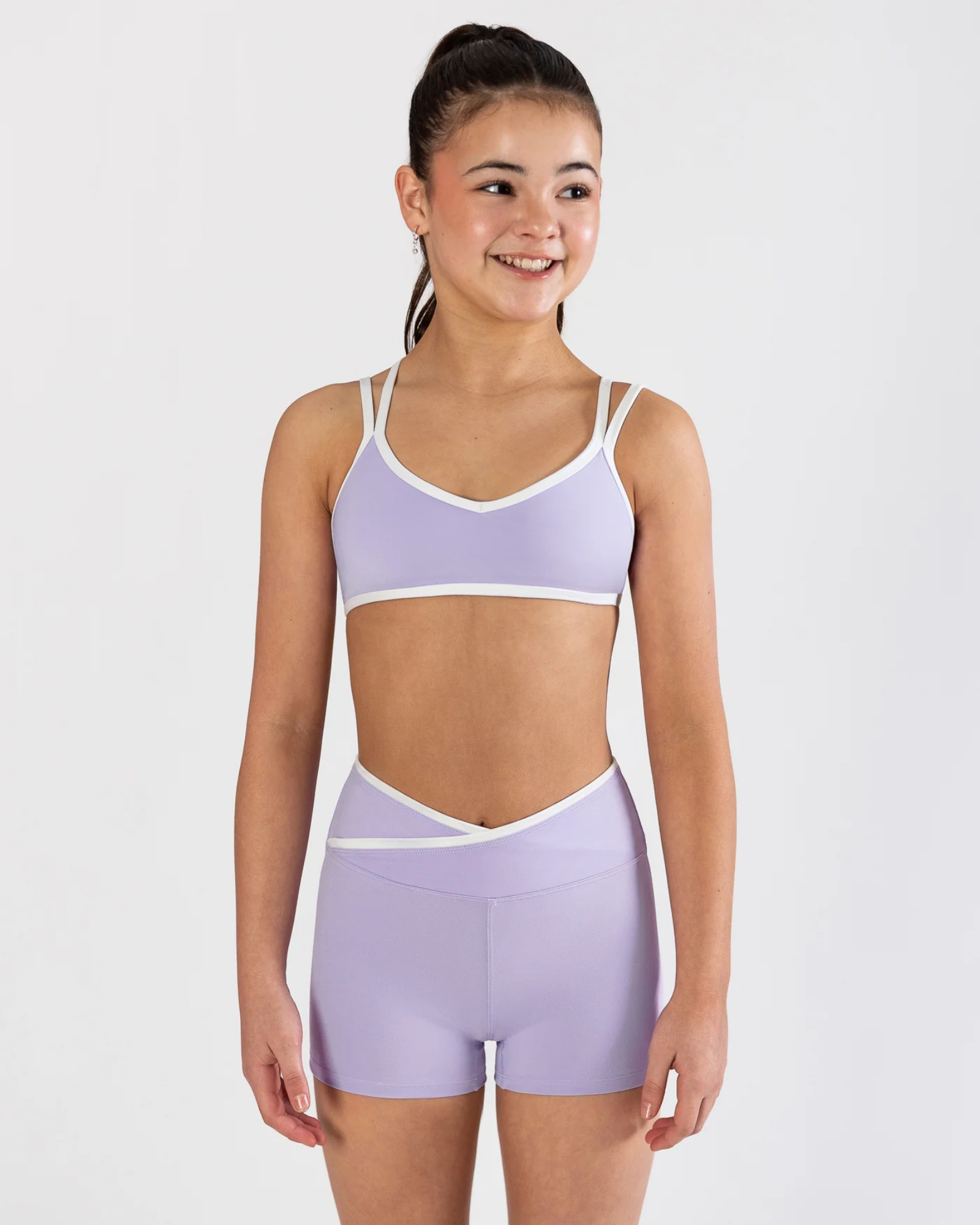 A young girl modeling a lavender athletic two-piece set consisting of a dance bra and matching high-waisted shorts with white trim, set against a plain light gray background.