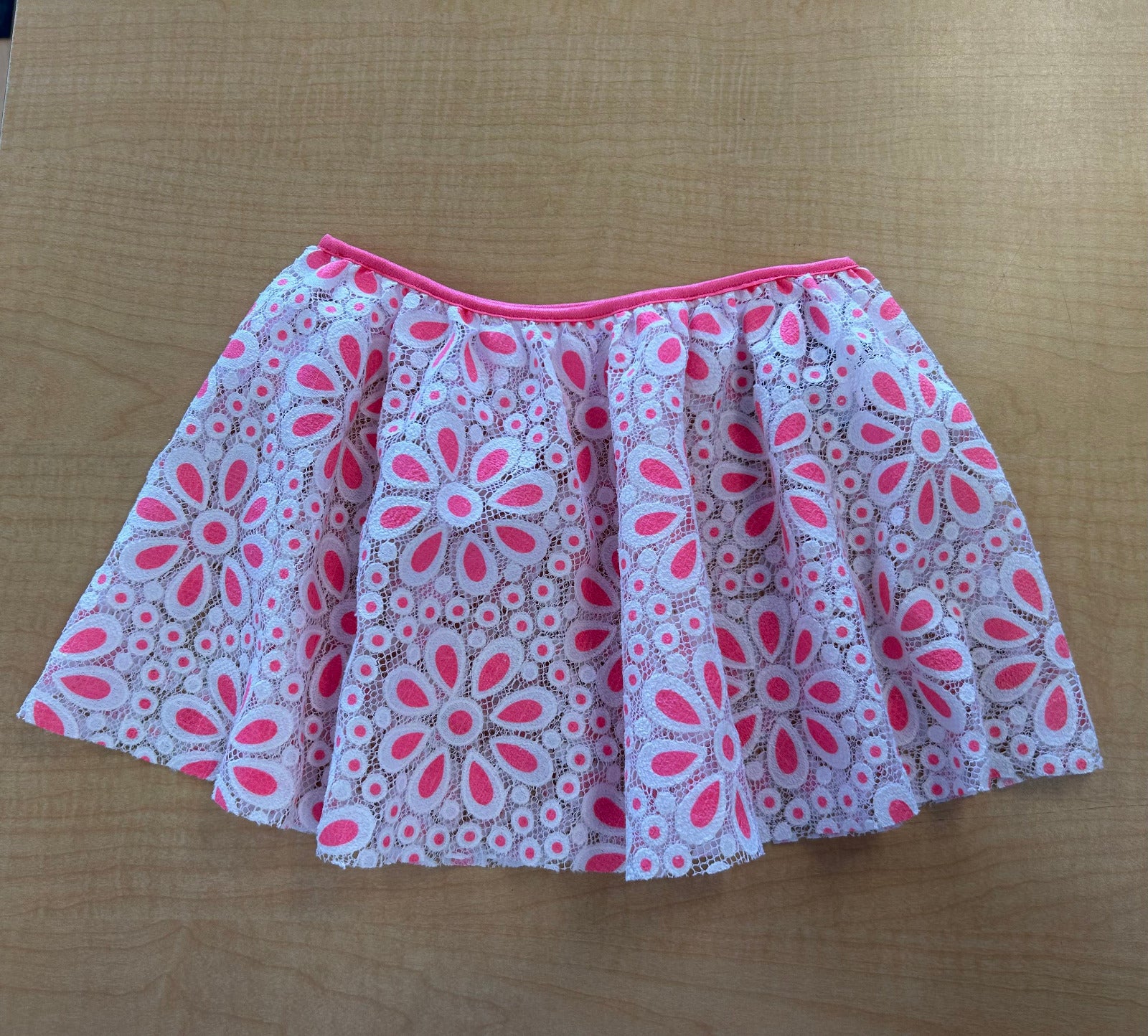 A short pleated skirt with a pink and white floral lace-like pattern, laid flat on a light brown wooden surface.