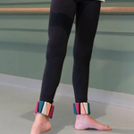 A person wearing black leggings and colorful ankle weights, standing barefoot in a studio with a green wall and ballet barre visible.