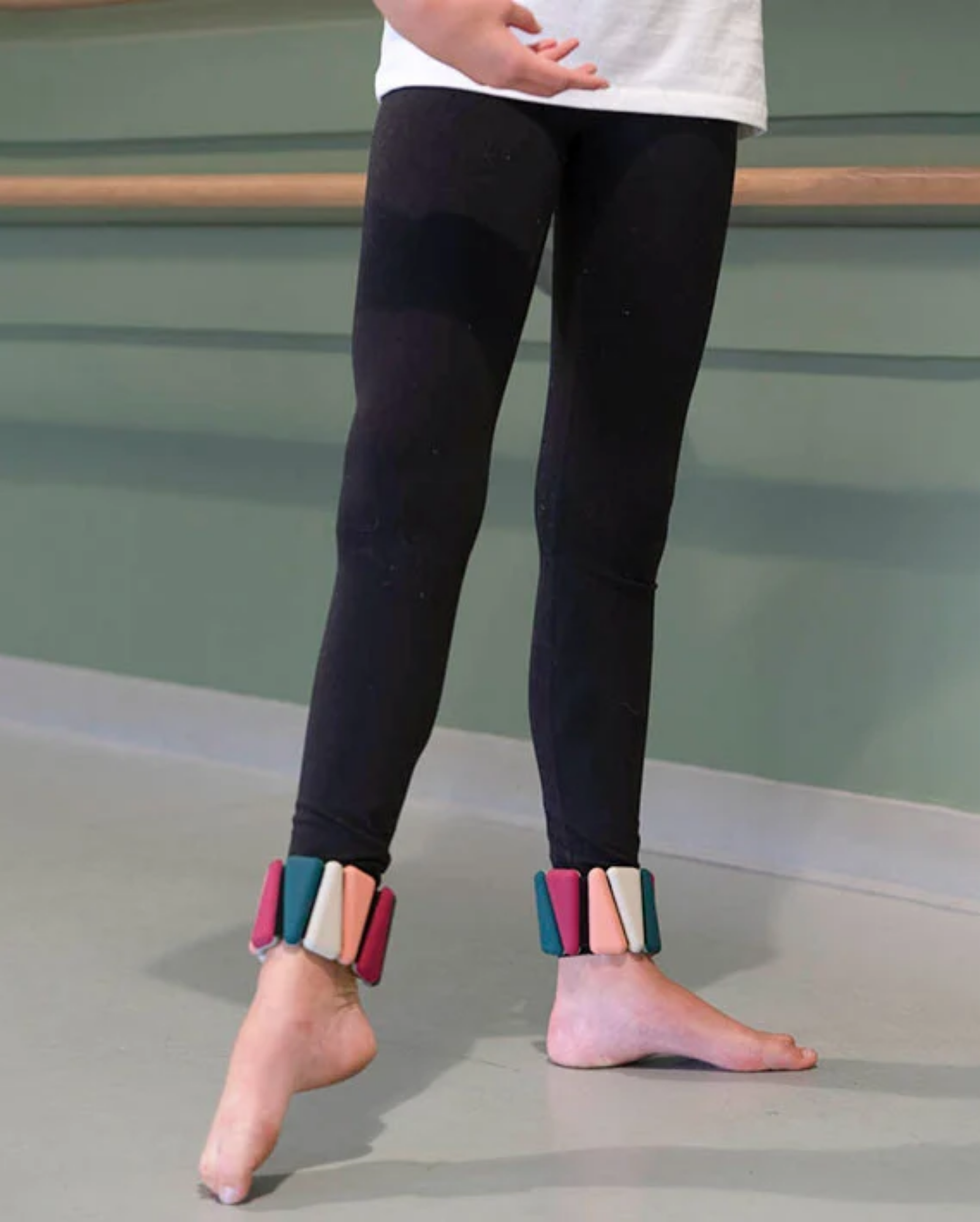 A person wearing black leggings and colorful ankle weights, standing barefoot in a studio with a green wall and ballet barre visible.
