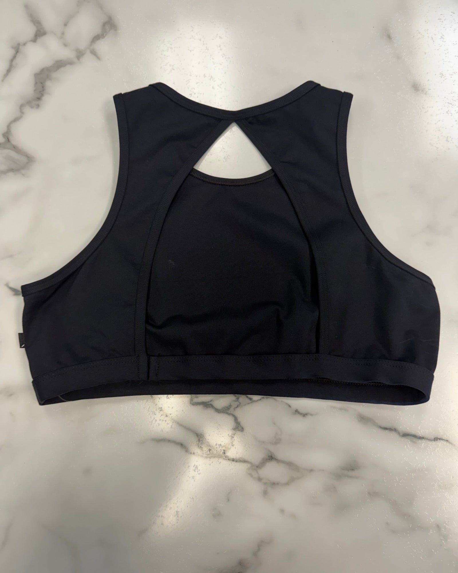 Black dance bra top shown from the back, featuring a crossover back design and wide underbust band, laid flat on a white marble surface.