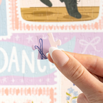 A close-up of a hand holding a small jigsaw puzzle piece in front of a colorful puzzle background with the word "DANCE" and illustrations of shoes and bows.
