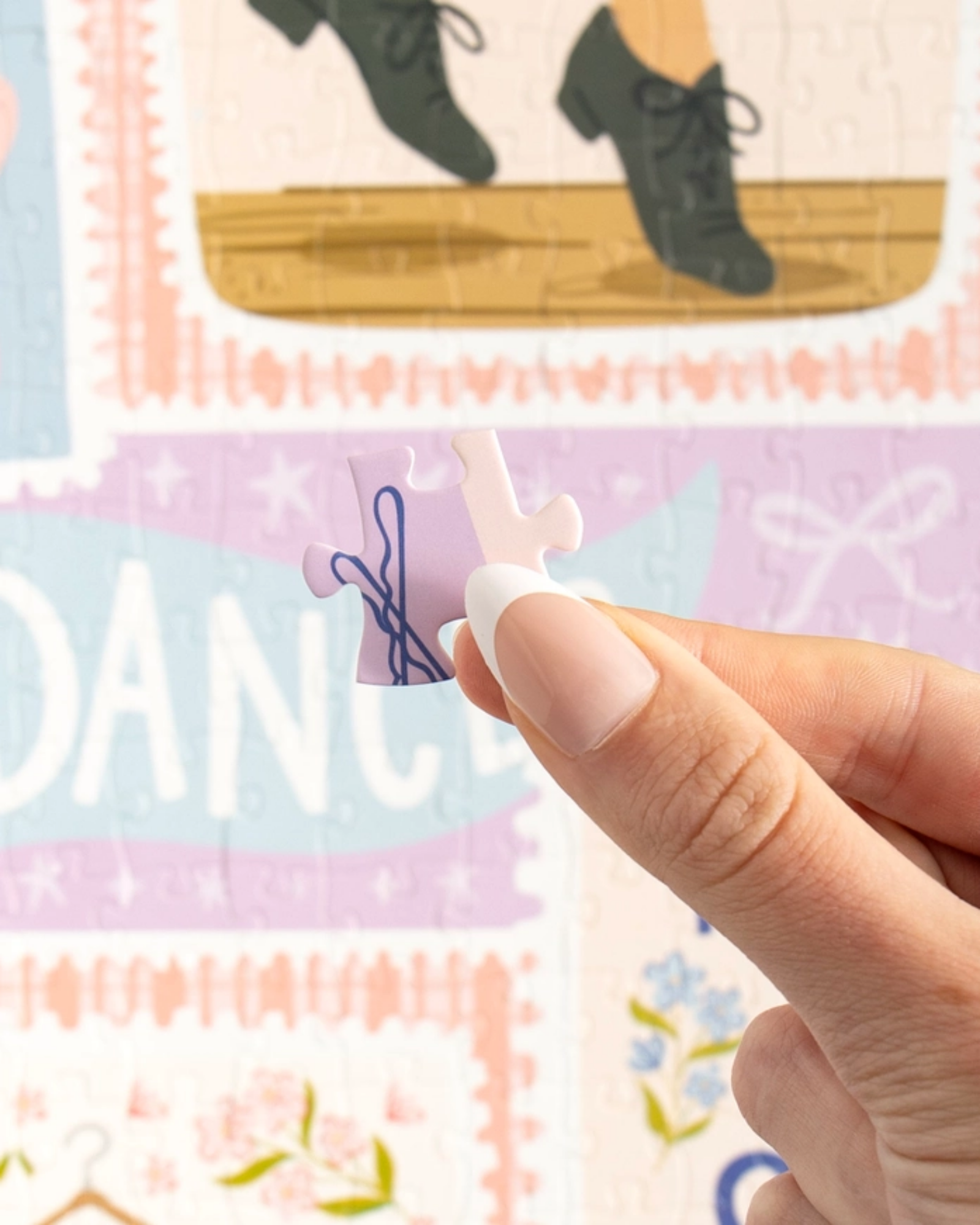 A close-up of a hand holding a small jigsaw puzzle piece in front of a colorful puzzle background with the word "DANCE" and illustrations of shoes and bows.