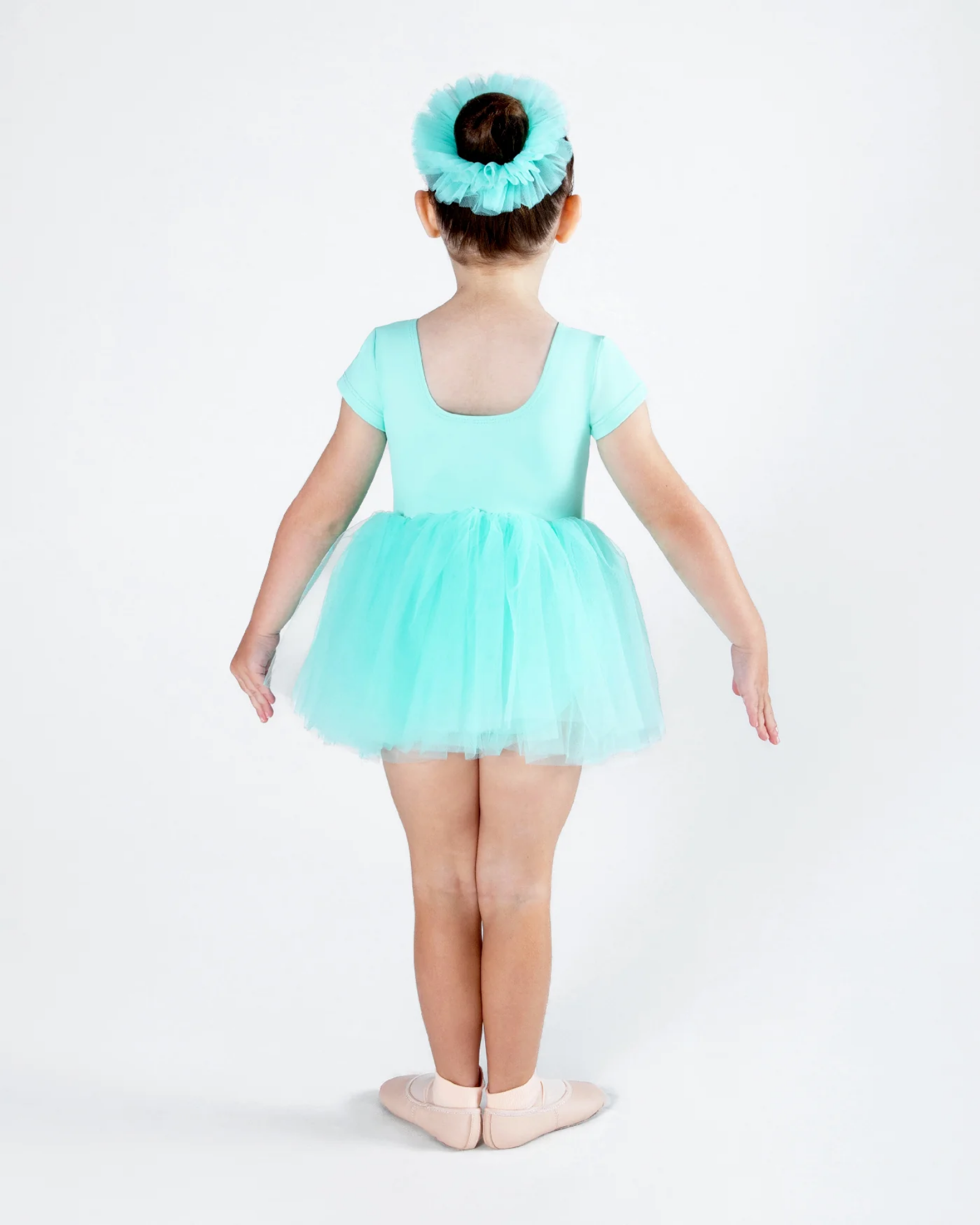 A young child viewed from the back wearing a mint green short-sleeve leotard with a matching tulle tutu skirt and a matching headband, standing on ballet slippers against a white studio background.