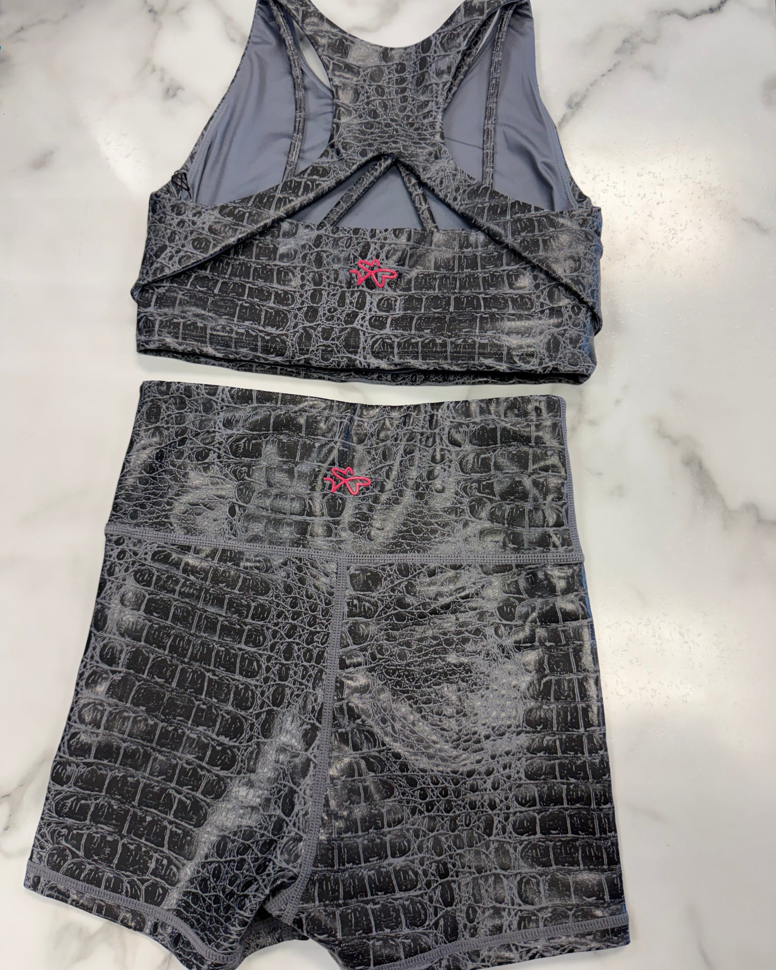 A two-piece activewear set consisting of a sports bra and matching shorts, both featuring a dark gray and black crocodile-embossed pattern with a small pink logo on the front. The set is laid flat on a white marble background.