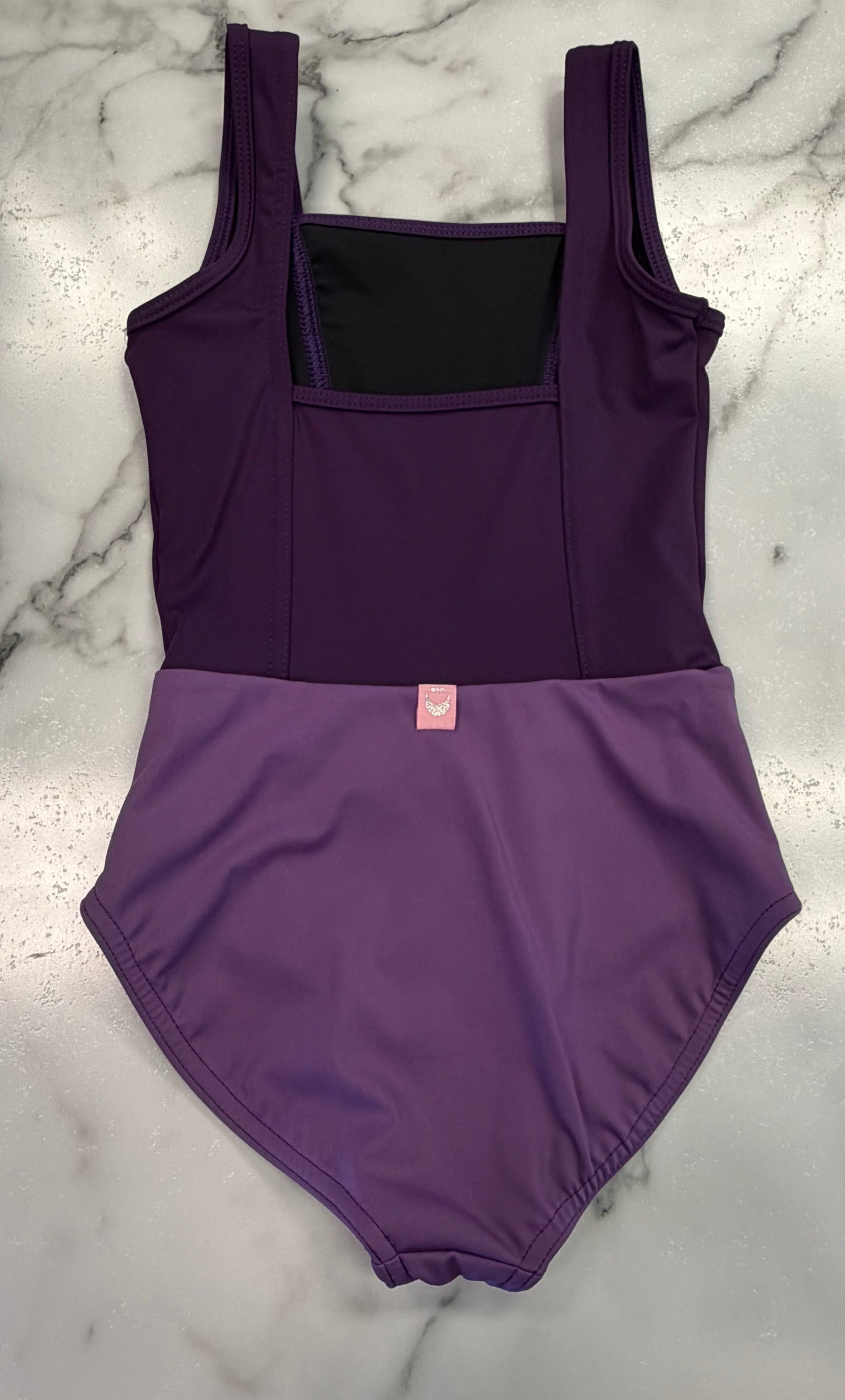 A two-tone purple leotard laid flat on a white and gray marble surface. The top portion is a dark purple, while the bottom is a lighter medium purple.