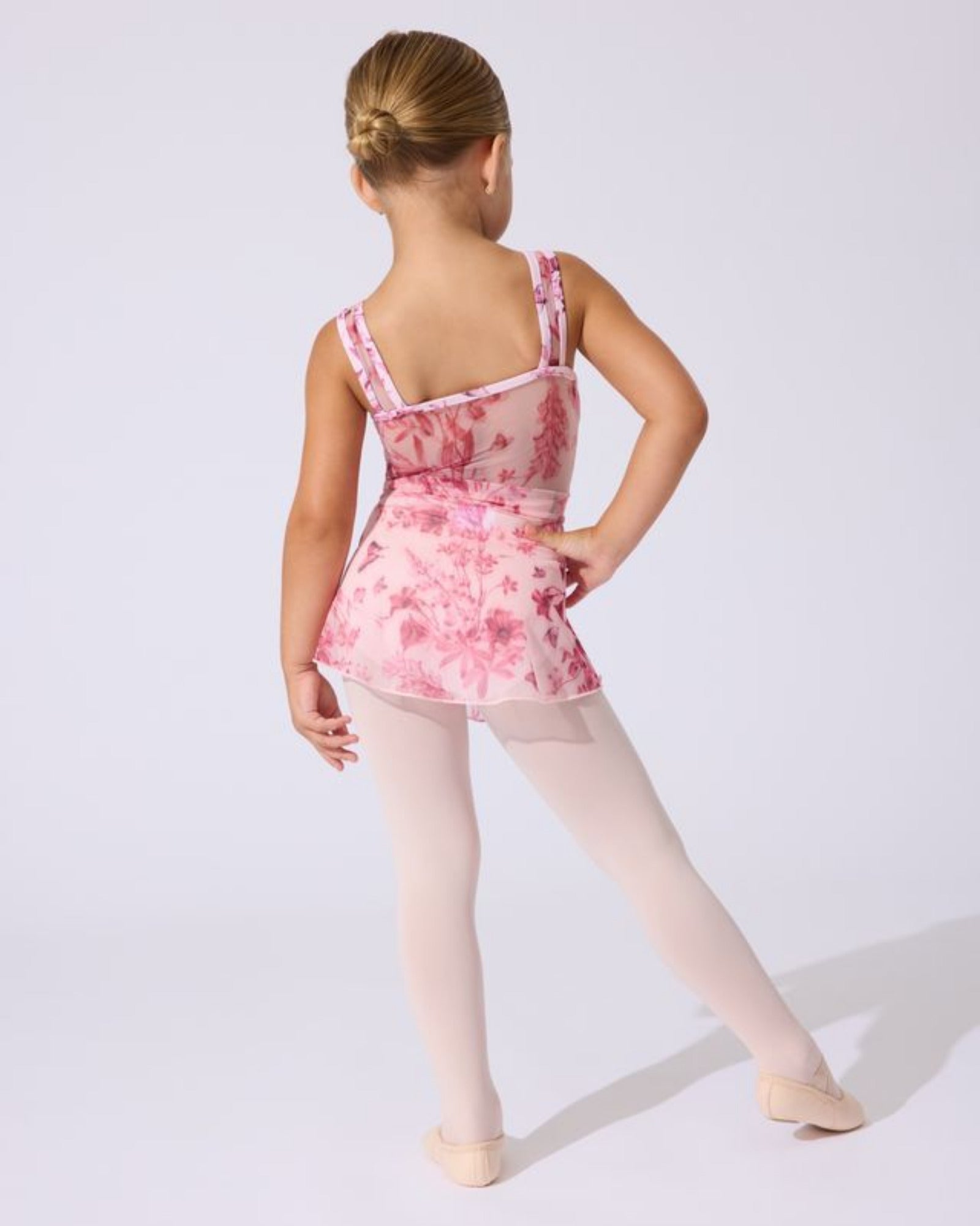 A young girl dancer shown from the back wearing a pink floral leotard, light pink tights, and ballet shoes against a plain light gray studio background.