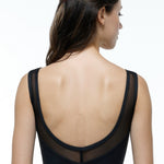 A woman shown from the back wearing a black sleeveless leotard with a sheer mesh back panel and a deep open back, photographed against a clean white studio background.