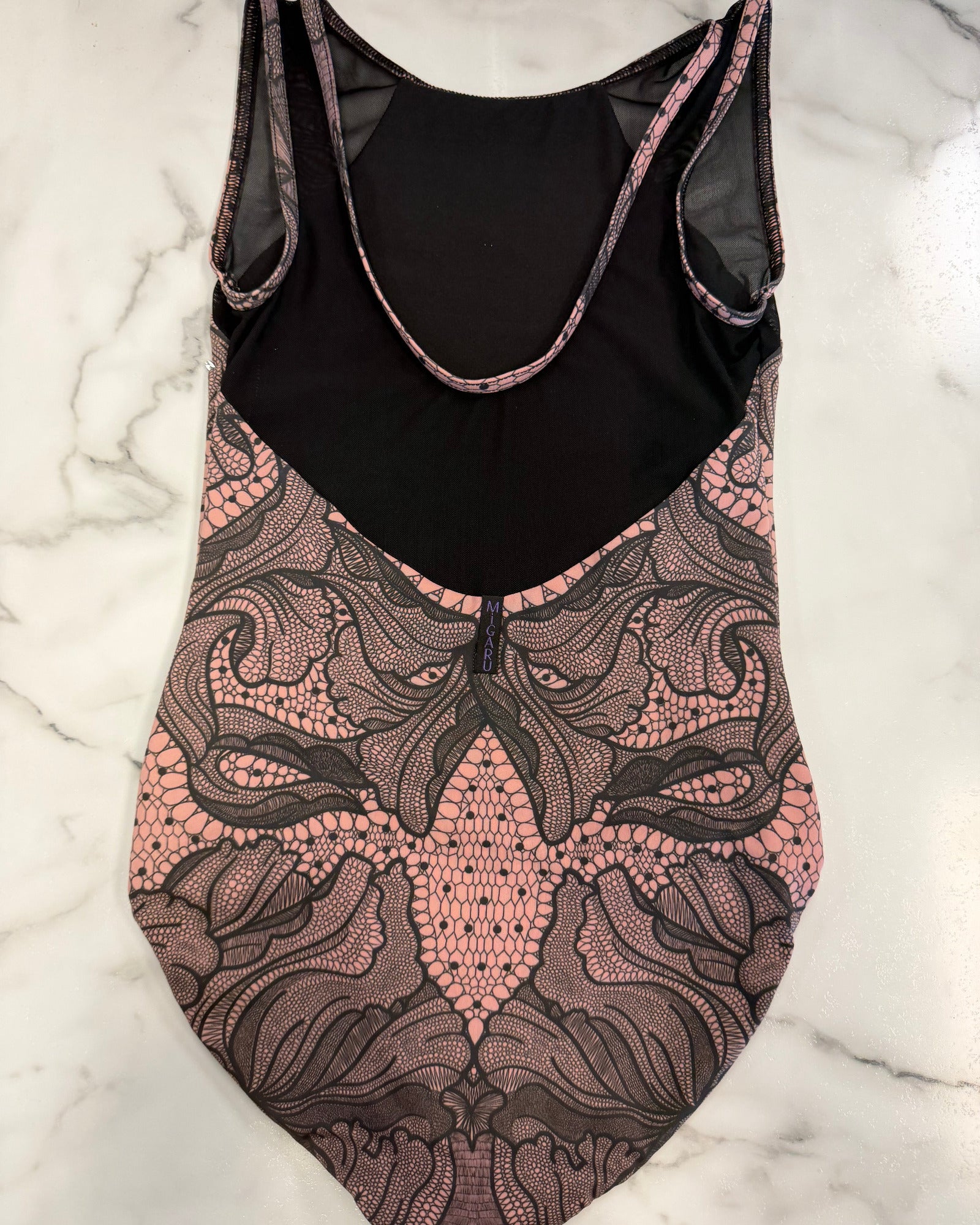 A sleeveless leotard laid flat on a white marble surface, featuring a black mesh upper back and black lining with a pink and black lace-like patterned lower body.