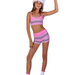 A young girl modeling a pink two-piece athletic set with a geometric pattern, including a sports bra and matching shorts, along with white socks and a pink and white fuzzy Santa hat, isolated on a white background.