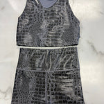 A two-piece activewear set featuring a sleeveless tank top and matching high-waisted shorts, both in a gray and black crocodile-embossed pattern, laid flat on a white marble background.