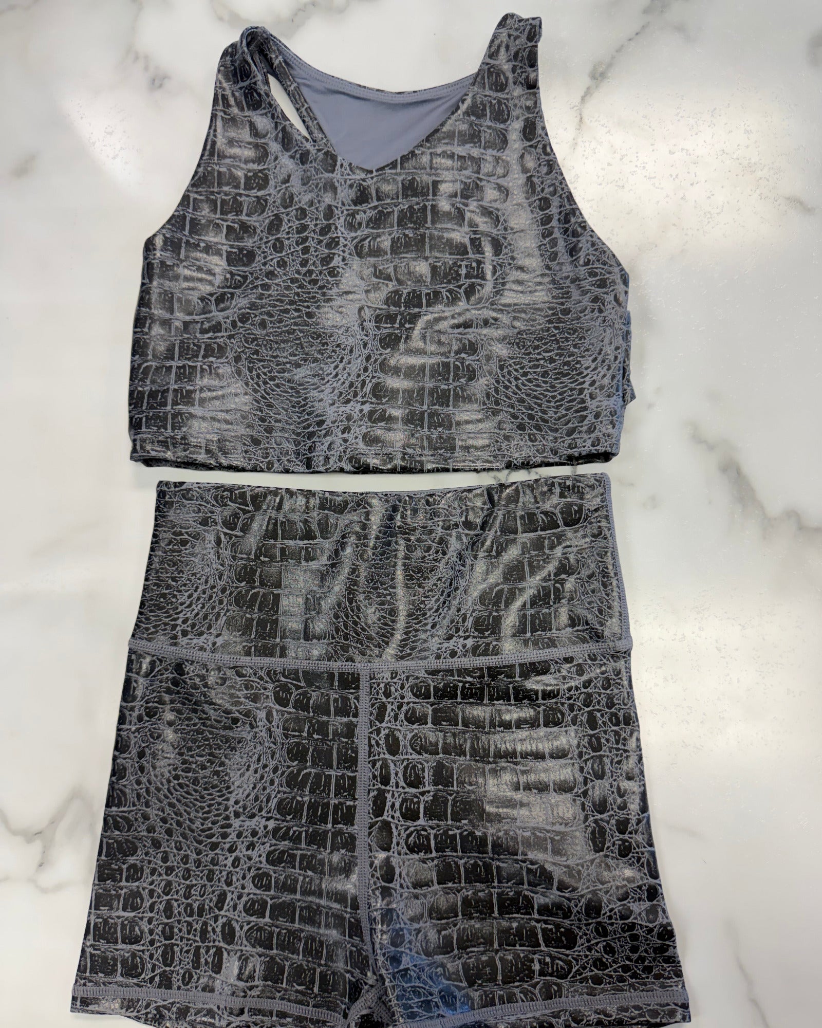 A two-piece activewear set featuring a sleeveless tank top and matching high-waisted shorts, both in a gray and black crocodile-embossed pattern, laid flat on a white marble background.