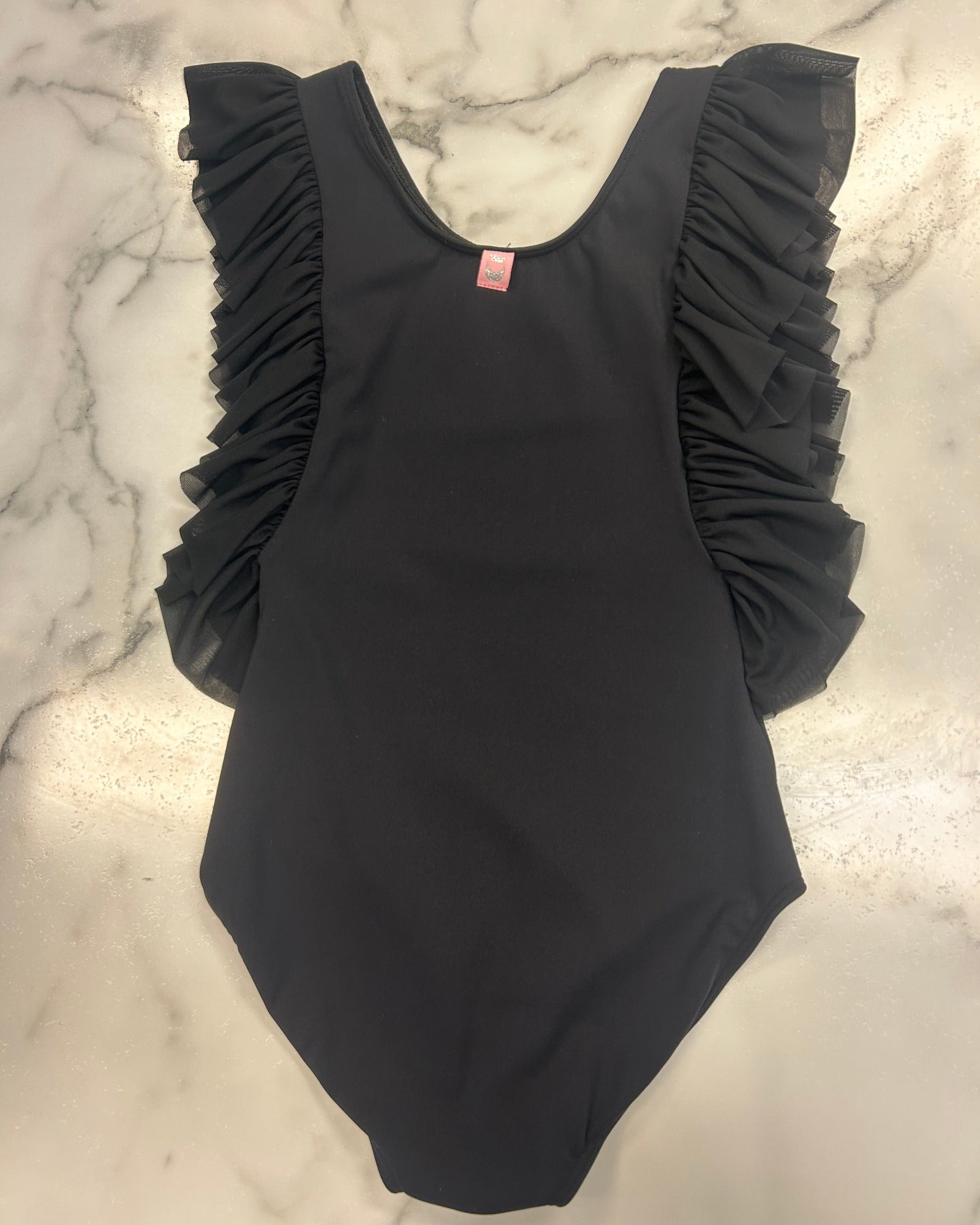 A black sleeveless leotard with dramatic ruffled shoulder details, laid flat on a white and gray marble surface.