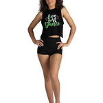 A woman modeling a black sleeveless crop top and matching black shorts against a plain white background. The top features a graphic print that reads 'live love dance' in white and green script.