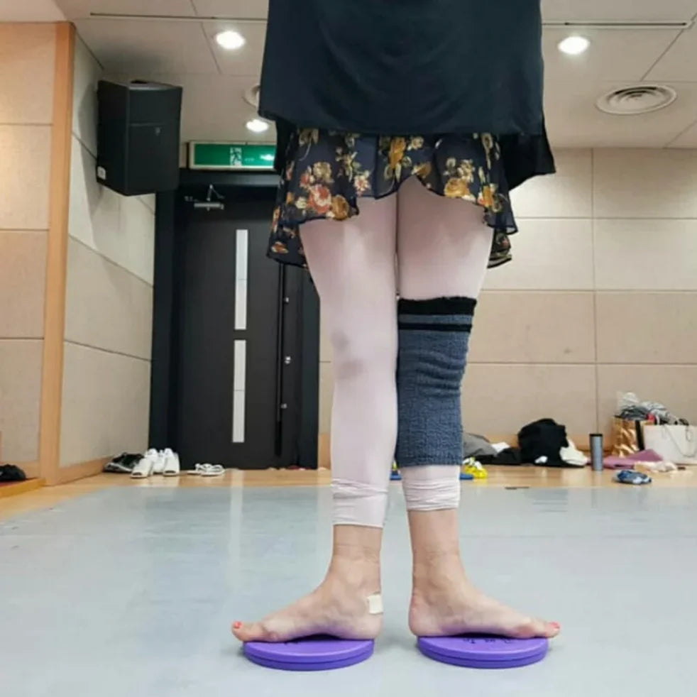 A person standing indoors on two purple balance discs, shown from the waist down. The person wears a black top, a floral skirt, light pink leggings, and a dark gray knee sleeve.