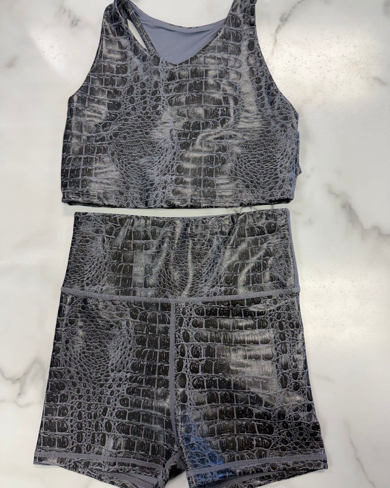 A two-piece activewear set featuring a sleeveless tank top and matching high-waisted shorts, both in a gray and black crocodile-embossed pattern, laid flat on a white marble background.